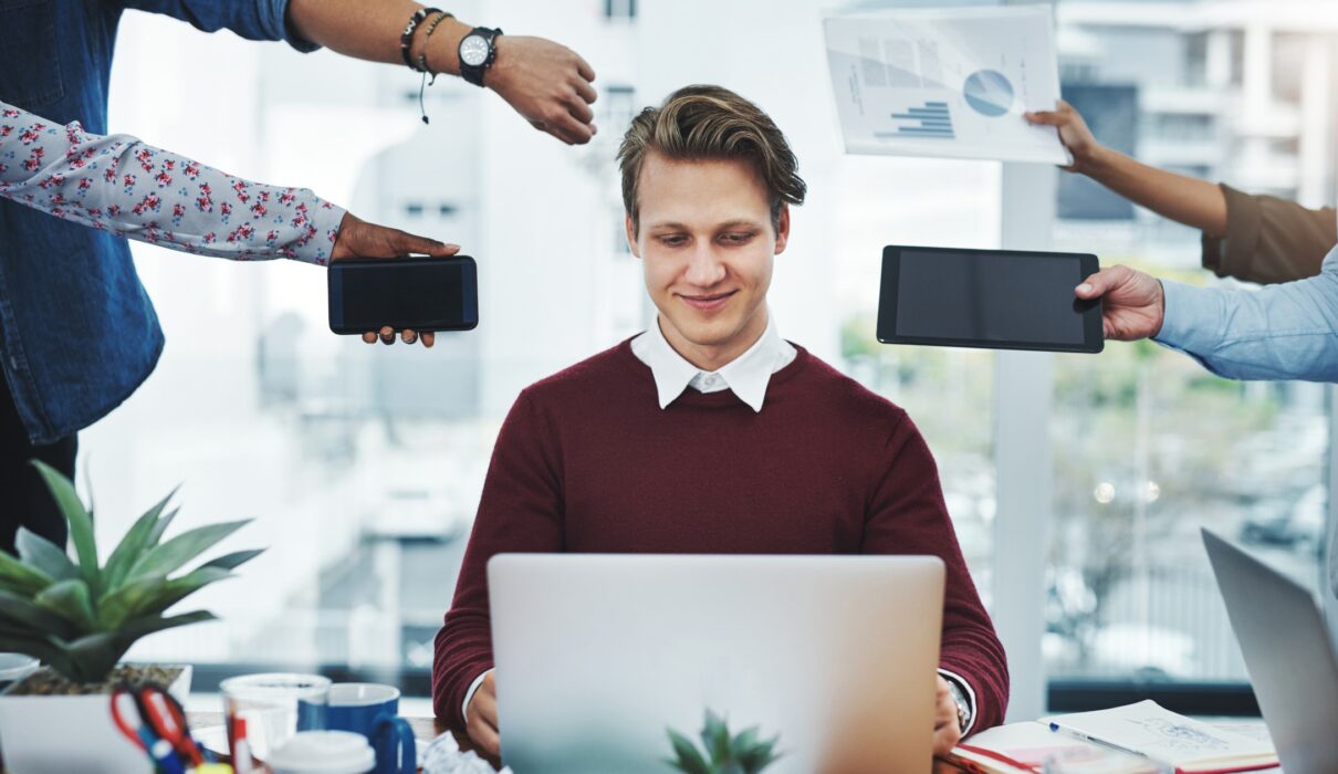 A man sitting calmly at a desk with a laptop, surrounded by multiple people holding smartphones, tablets, and charts, illustrating the concept of staying composed and focused despite numerous distractions and demands.